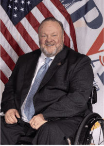 A smiling man in a suit and tie sits in a wheelchair in front of an American flag and a colorful banner.